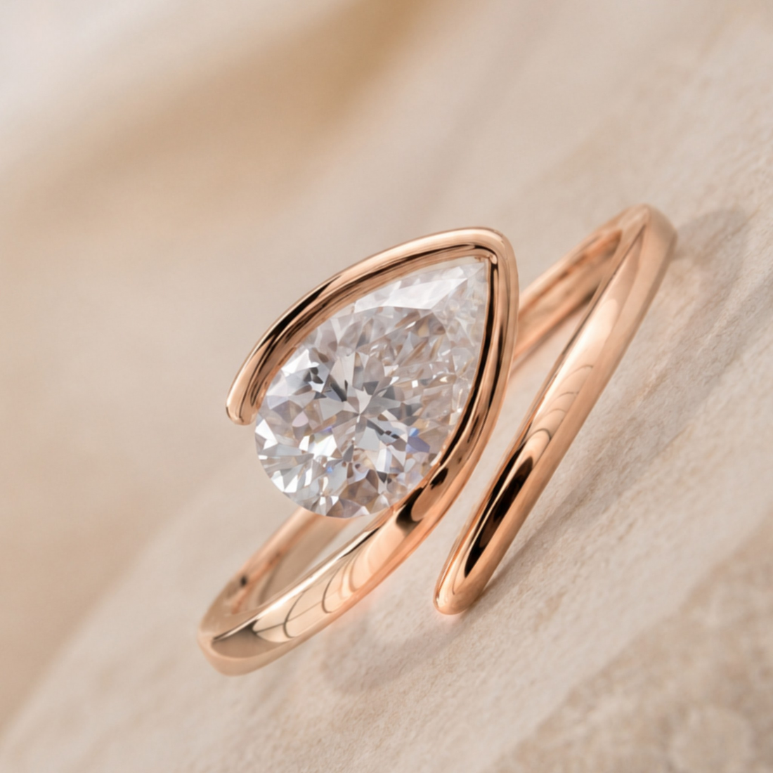 A rose gold ring featuring a large pear-cut clear gemstone in a curved setting, resting on a soft beige textured surface.