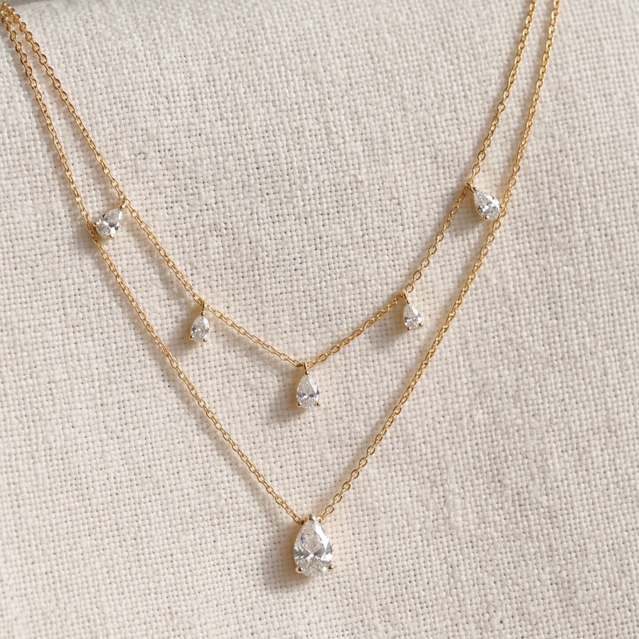 A delicate double-layered gold necklace with small clear teardrop gemstones and a larger pear-shaped clear stone pendant, displayed on a textured beige fabric background.