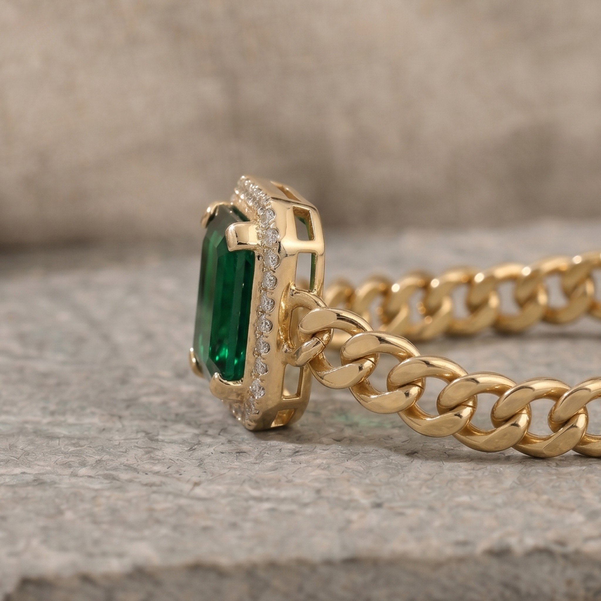 A gold chain necklace with a rectangular green emerald-cut center stone and a halo of small clear stones, resting on a textured gray stone surface.