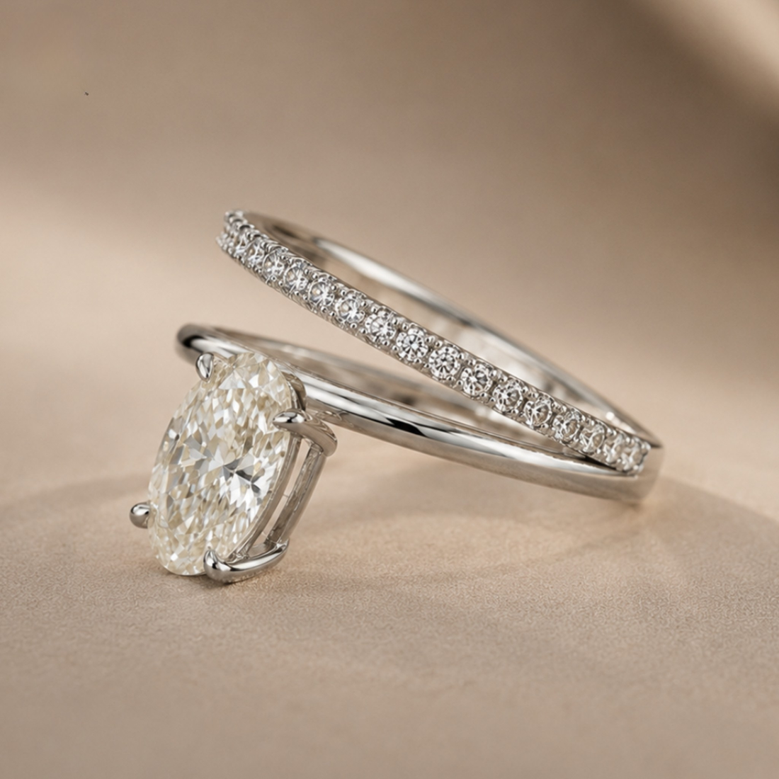 A silver engagement ring with a large oval-cut diamond and a matching pavé band ring, displayed on a soft beige background.