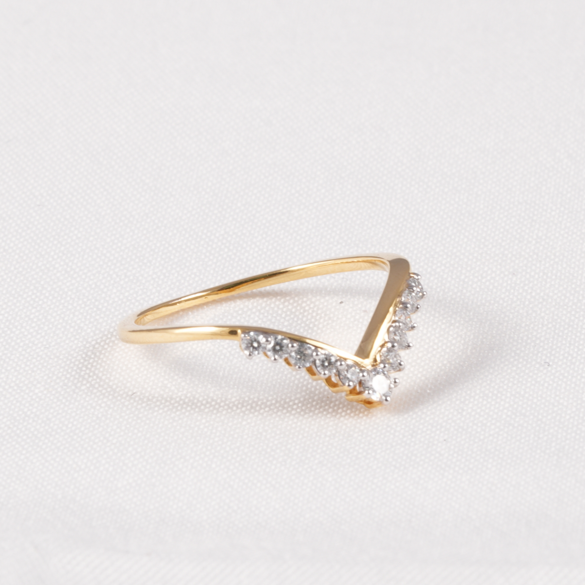 A gold ring with a V-shaped design, featuring a row of small clear stones along the front curve, set against a textured white background.