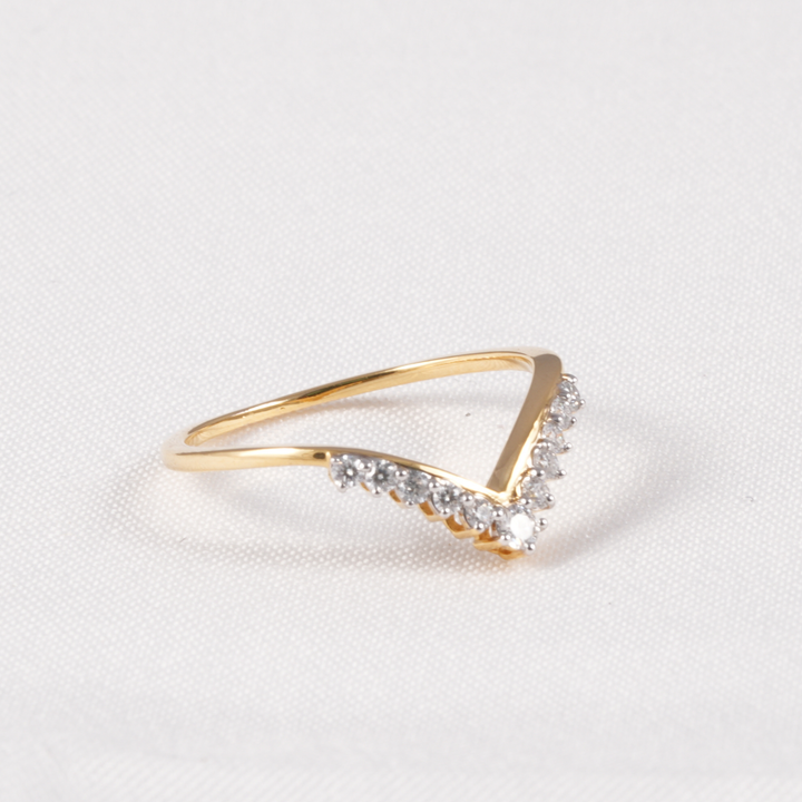 A gold ring with a V-shaped design, featuring a row of small clear stones along the front curve, set against a textured white background.