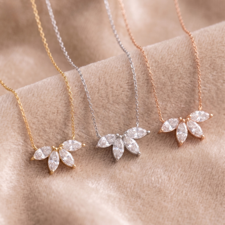 Three delicate flower-shaped pendant necklaces with marquise-cut clear stones, shown in gold, silver, and rose gold tones on a soft beige fabric background.