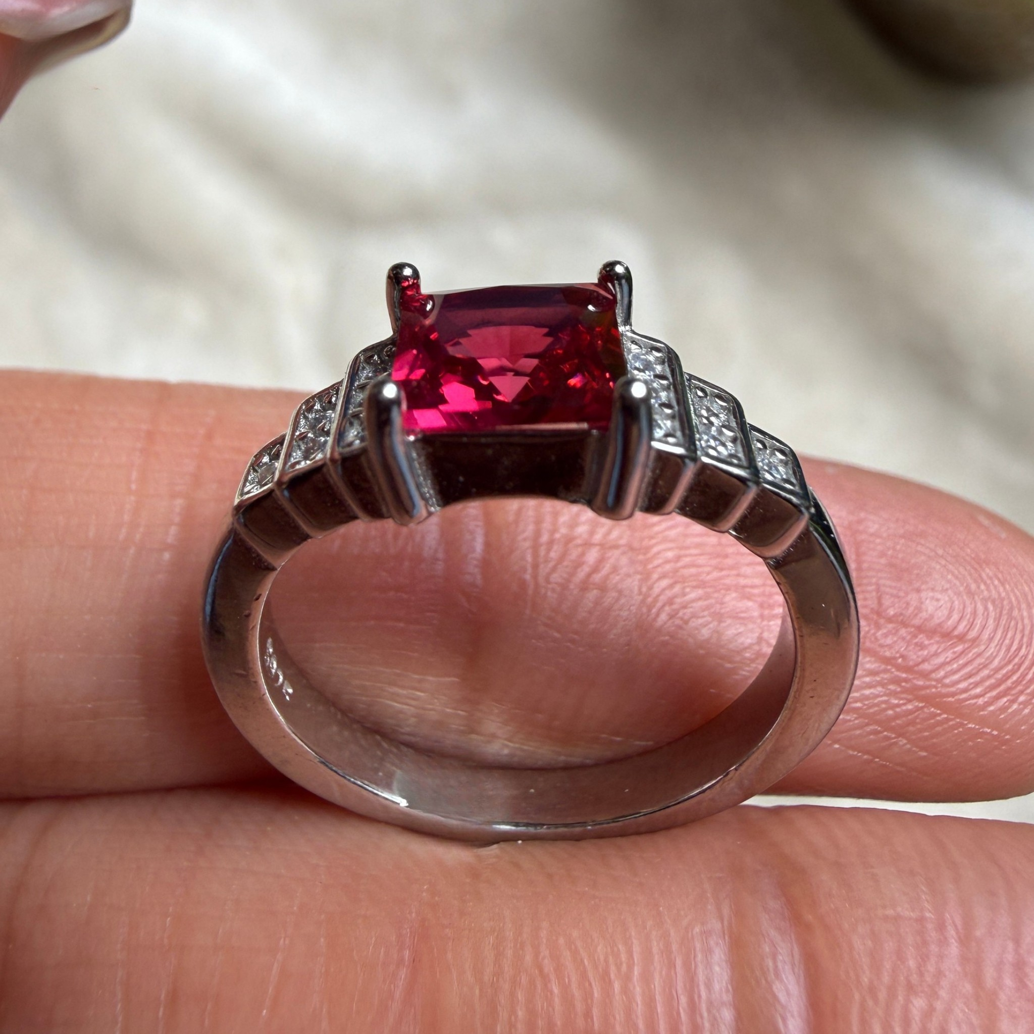 A silver ring with a rectangular red gemstone and small clear accent stones, shown resting on a person's fingers against a soft beige fabric background.