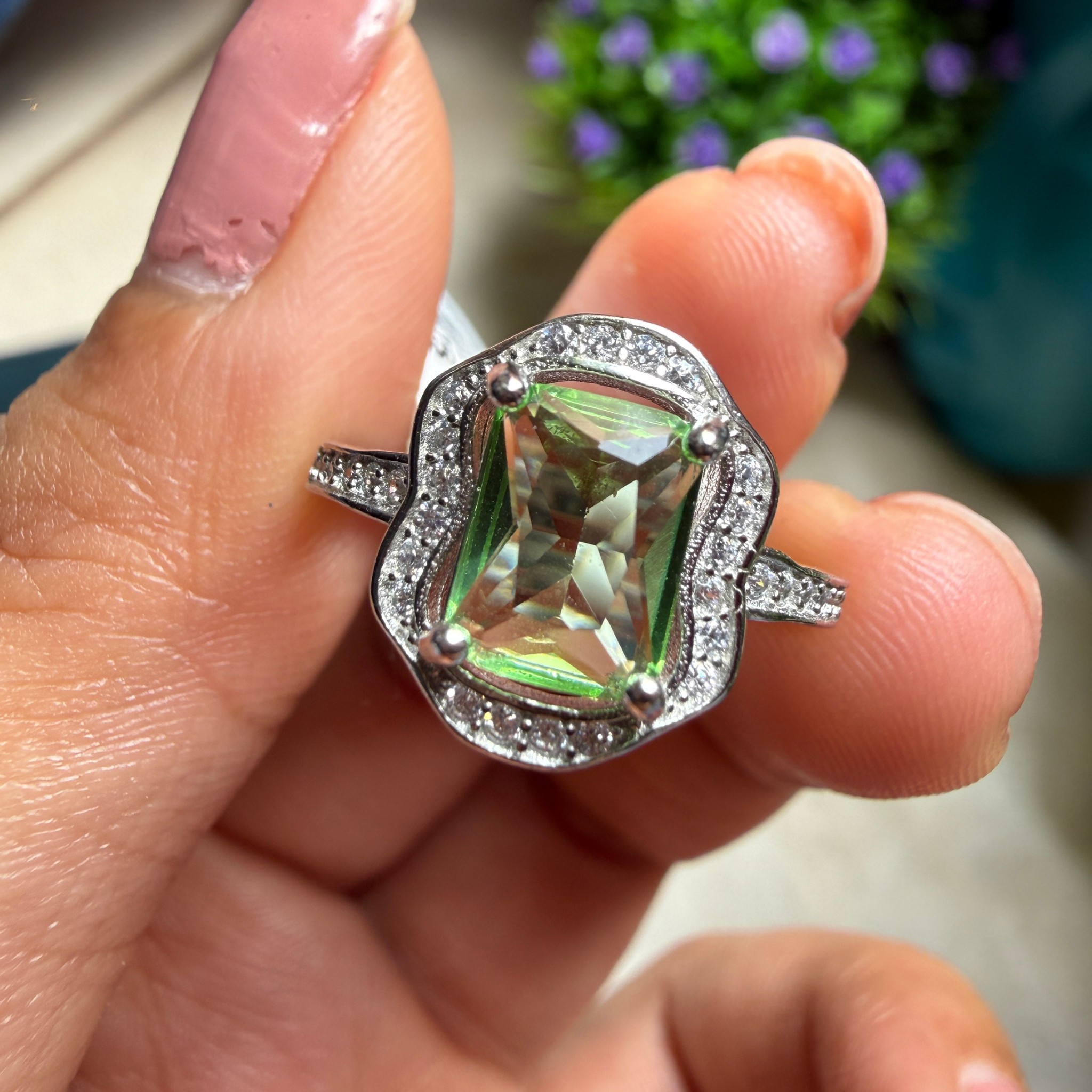 A close-up of a hand holding a silver ring with a large iridescent green rectangular gemstone surrounded by a halo of small clear stones.