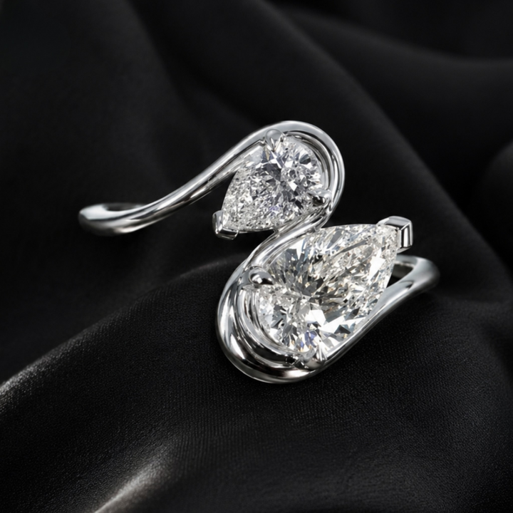 A silver-toned ring featuring two large pear-cut clear gemstones in a swirling open-band design, photographed on draped black fabric.