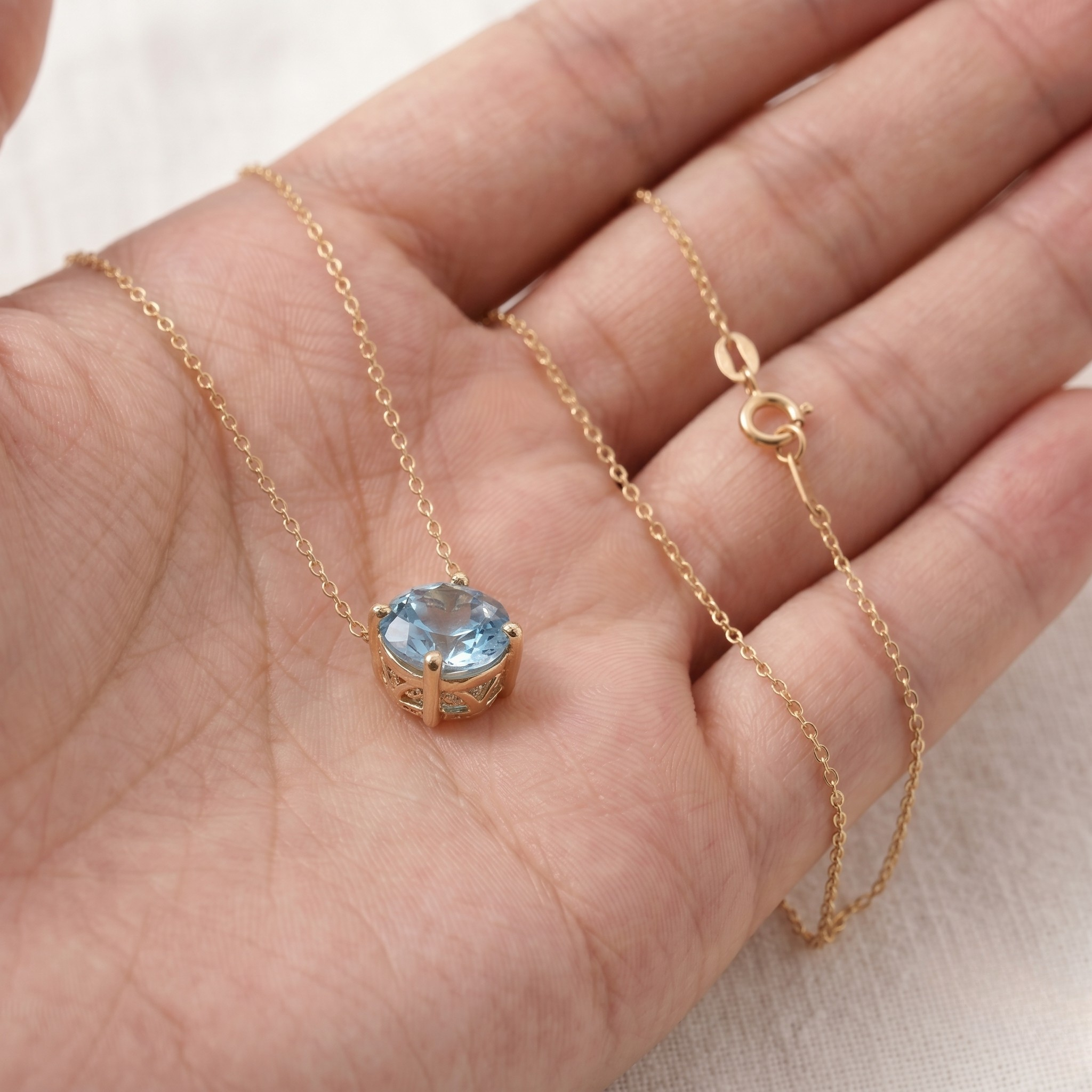 A delicate gold chain necklace with a small round bezel-set light blue gemstone pendant, displayed on an open hand.