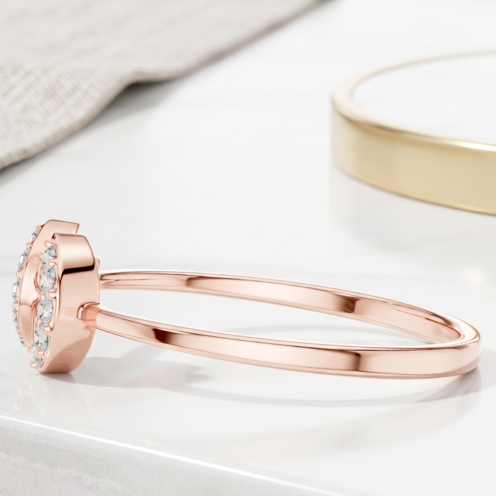 A rose gold ring with a bezel-set diamond cluster, shown in a close-up product shot on a white marble surface with a neutral beige fabric and a gold bangle in the background.