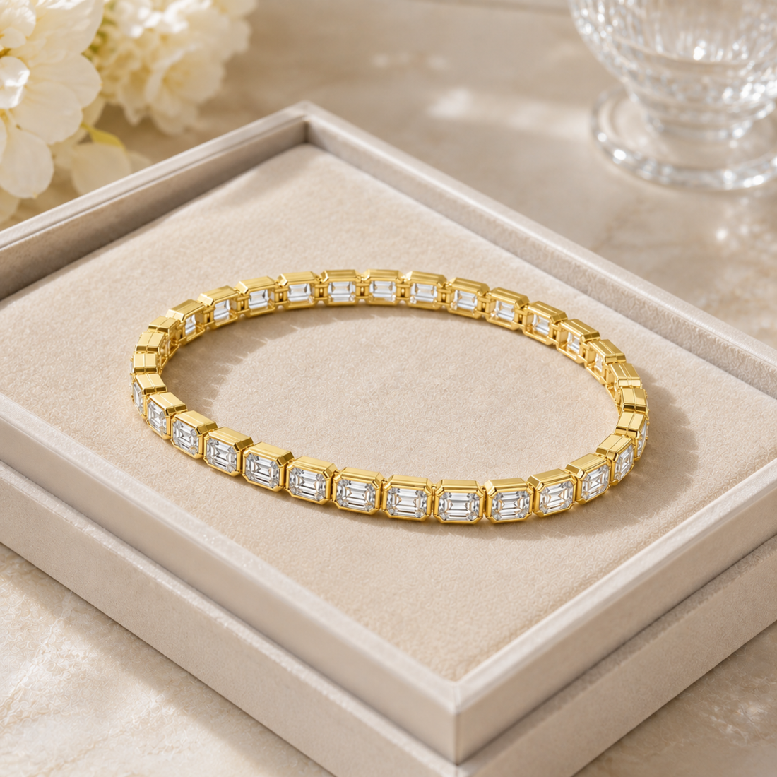 A gold tennis bracelet with rectangular Emerald -cut clear stones, displayed on a beige jewelry box tray.