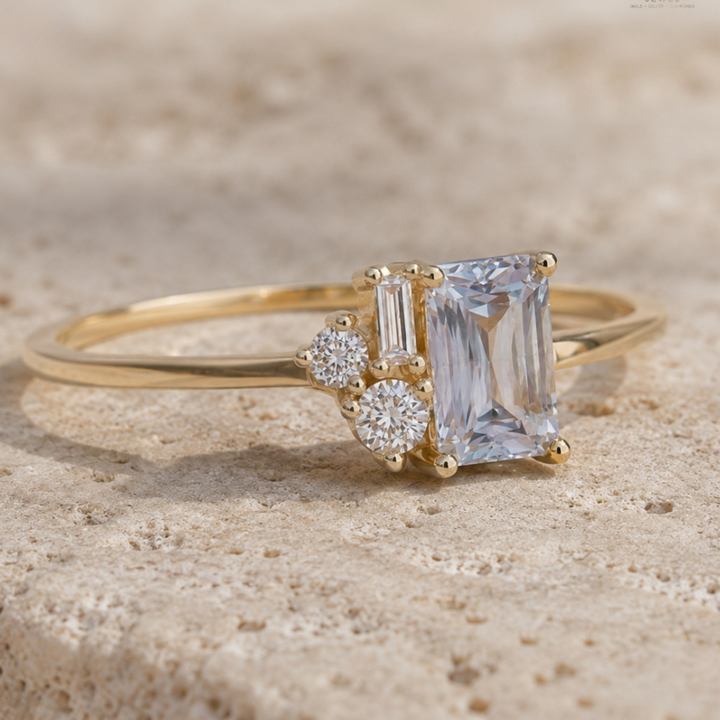 A yellow gold ring featuring a large emerald-cut clear center stone and two smaller clear accent stones, photographed on a textured beige stone surface.
