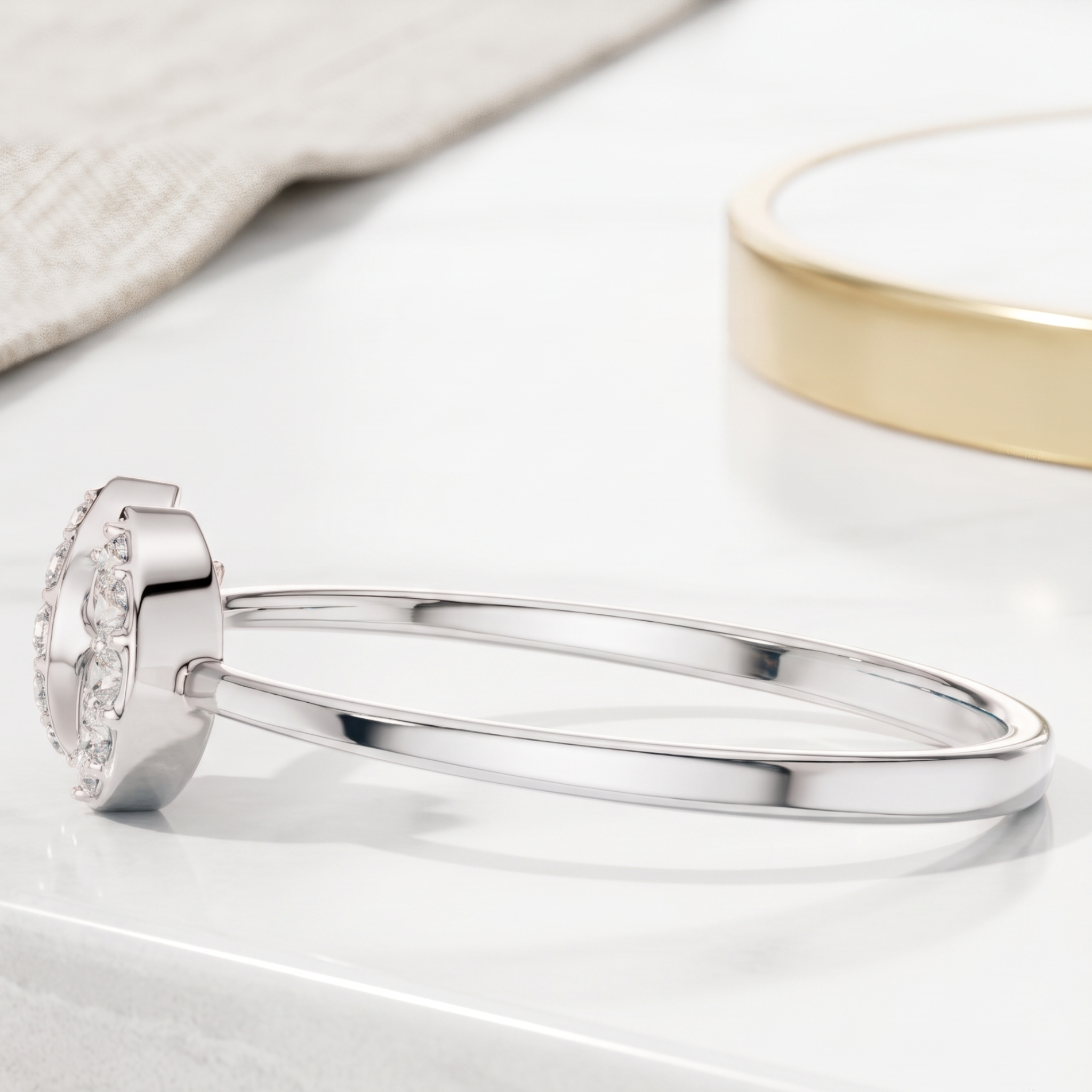 A polished silver-toned bangle bracelet with a decorative oval end set with small clear stones, displayed on a white surface with a soft beige fabric and a gold-toned circular object in the background.