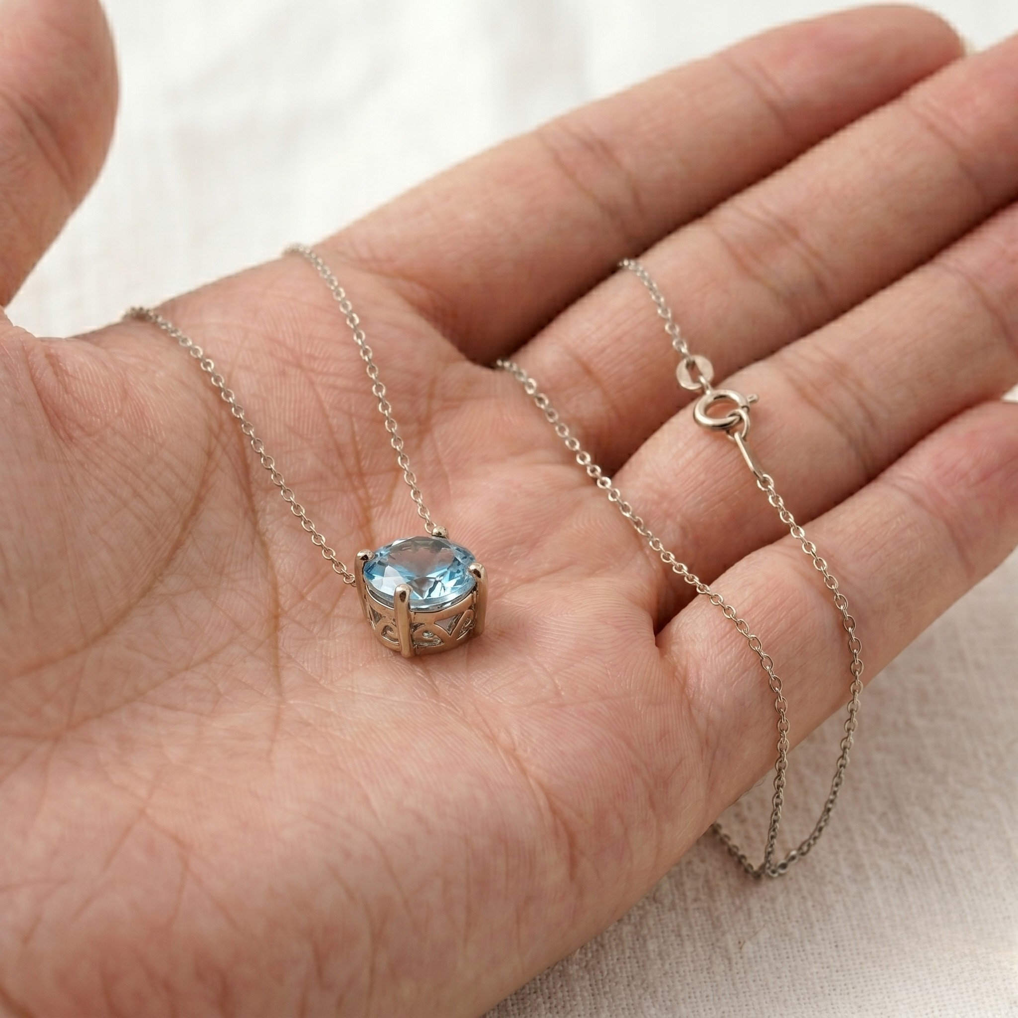 A silver necklace with a heart-shaped bezel-set light blue gemstone pendant, draped over an open hand against a soft white fabric background.