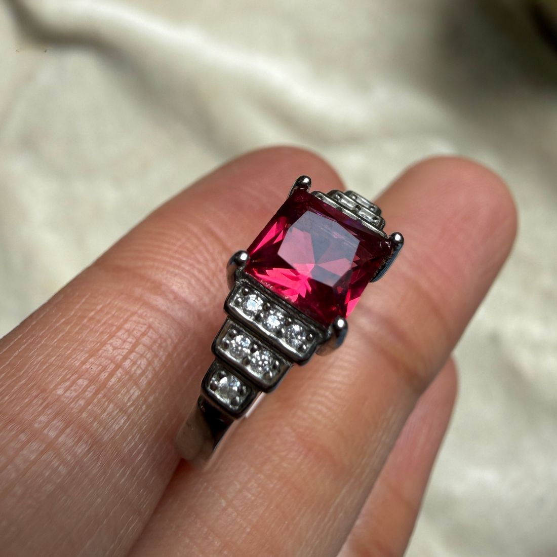 A close-up of a silver-toned ring with a large rectangular pink gemstone and small clear stones, shown worn on a person's fingers against a soft light fabric background.