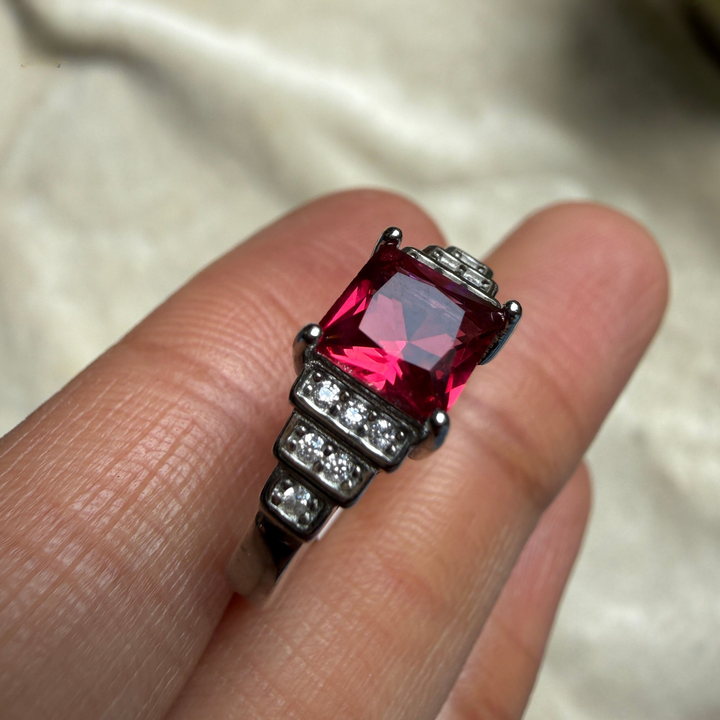 A close-up of a silver-toned ring with a large rectangular pink gemstone and small clear stones, shown worn on a person's fingers against a soft light fabric background.