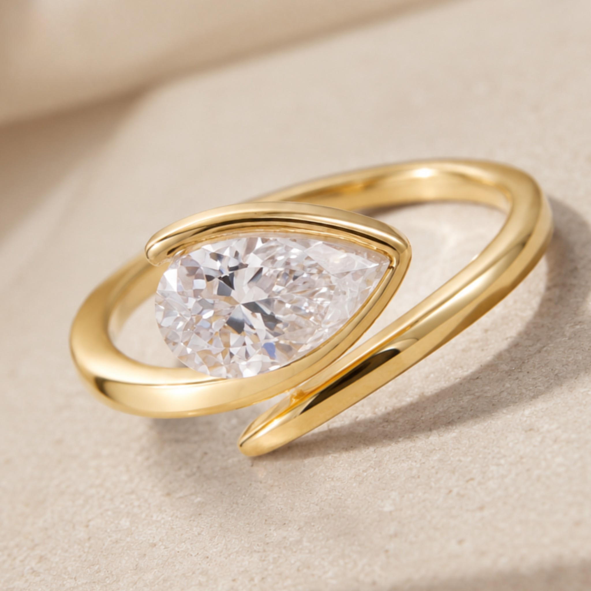 A gold ring with a teardrop-shaped clear gemstone, resting on a textured beige surface.
