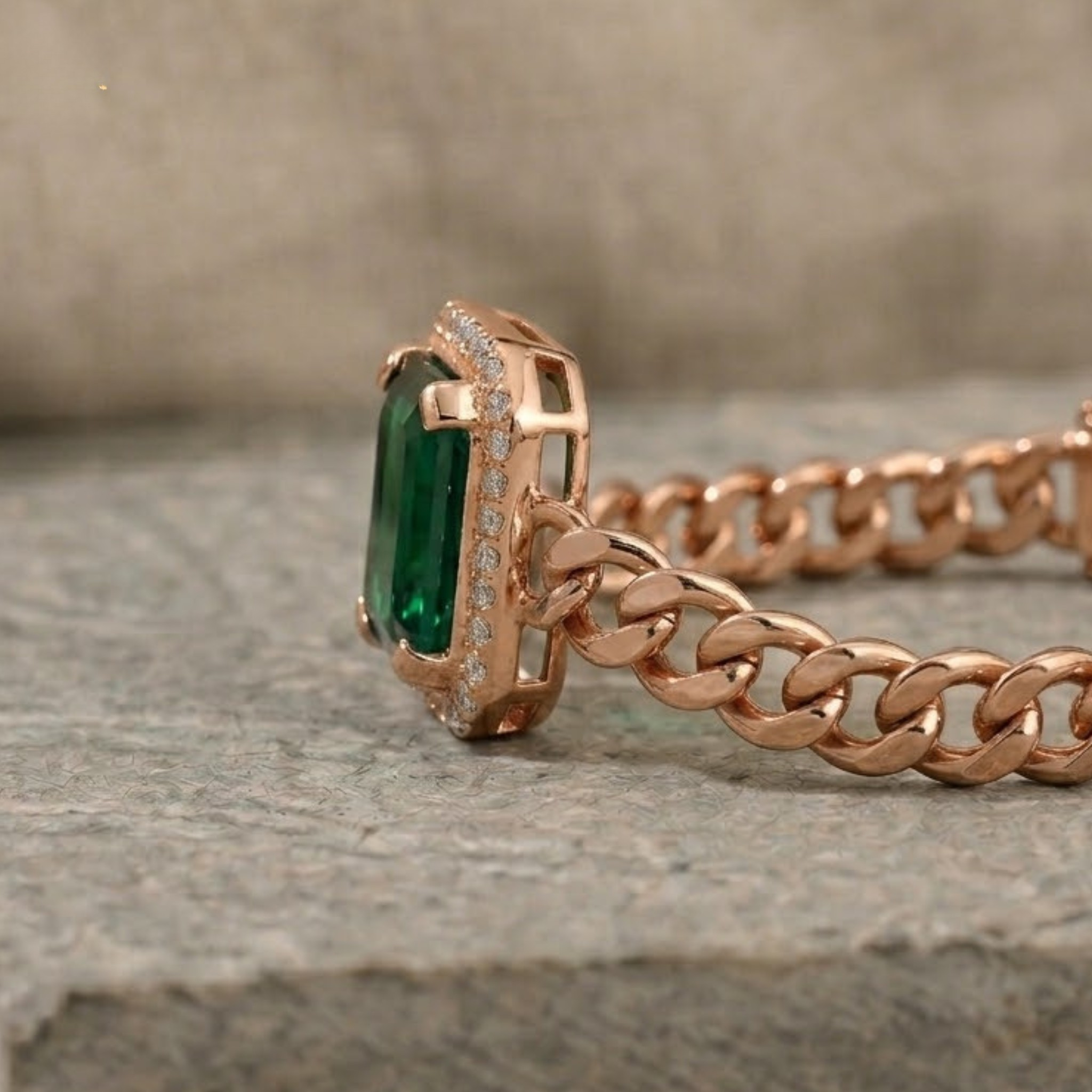 Rose gold chain ring with a rectangular green emerald-cut center stone and small clear accent stones, shown close up on a stone surface.