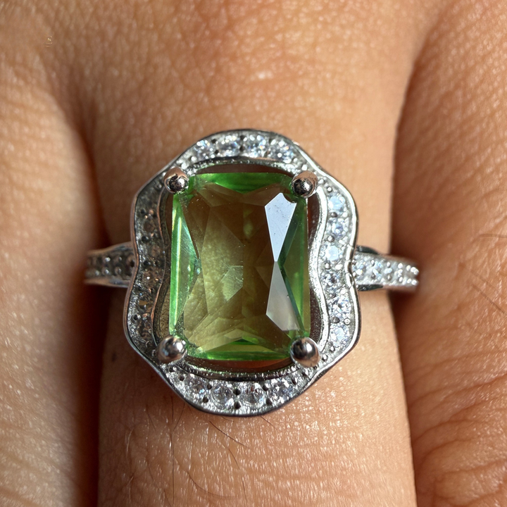 A close-up of a ring worn on a finger, featuring a large green rectangular gemstone in a silver-toned setting with small clear accent stones around the border and along the band.
