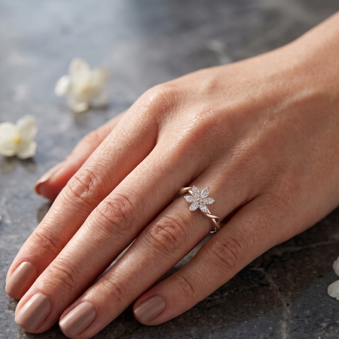 A close-up of a hand wearing a delicate rose gold ring shaped like a small flower with clear stones, resting on a dark textured surface with soft white flower petals in the background.