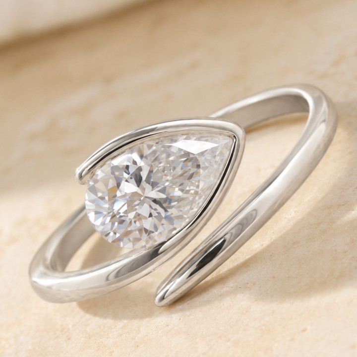 A silver-toned open band ring featuring a large, faceted pear-cut clear gemstone in a prong setting, resting on a light beige textured surface.