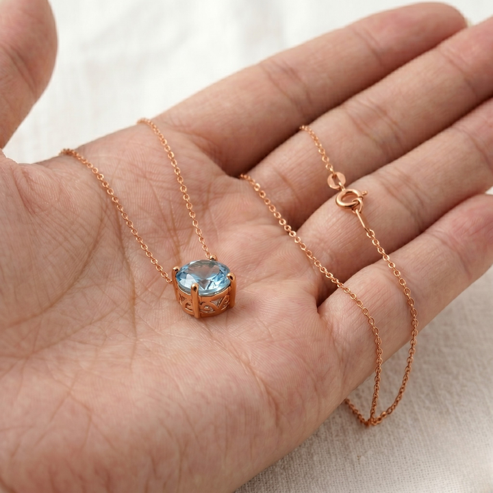 A rose gold necklace with a heart-shaped bezel-set light blue gemstone, shown draped over an open hand to demonstrate scale.