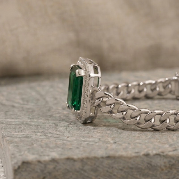 A close-up of a silver-toned Cuban link chain bracelet with a rectangular green emerald-cut center stone and a halo of small clear stones, resting on a textured gray stone surface.
