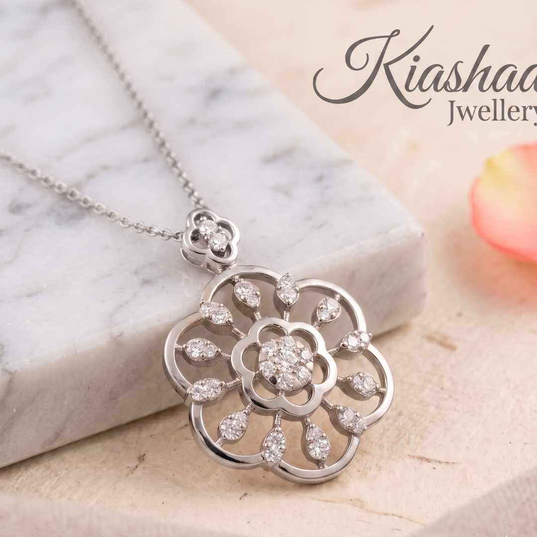 A silver-tone floral pendant necklace with clear stones, displayed on a white marble block over a light beige surface. The brand name 'Kiashaa Jewellery' is visible in the upper right.