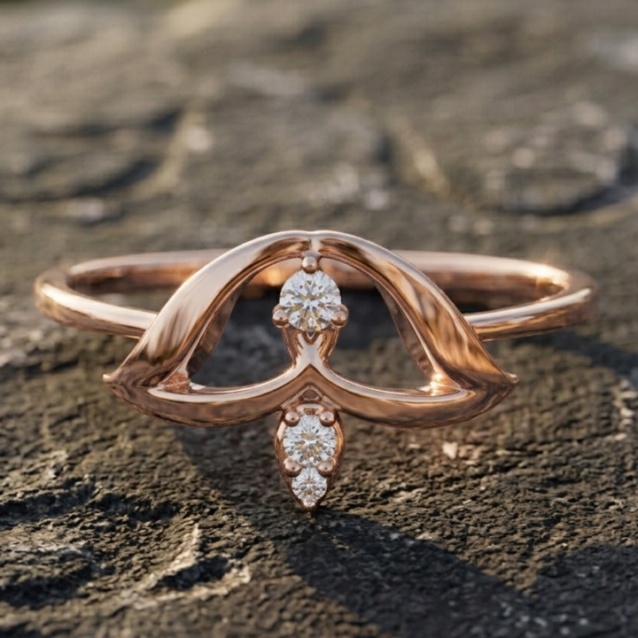 A rose gold ring with a sculptural, openwork design and small clear gemstones, resting on a textured dark stone surface.