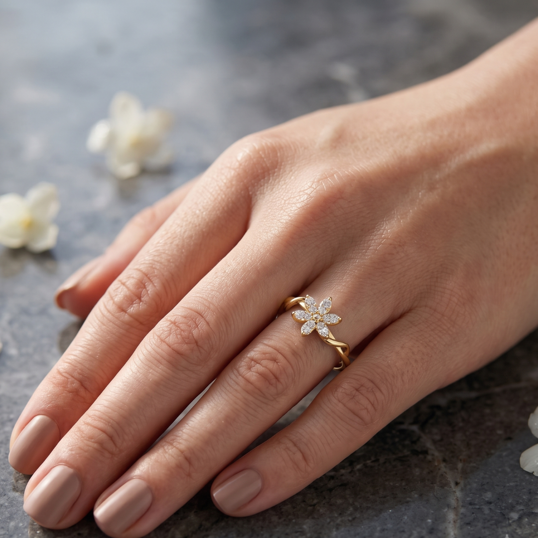 A close-up of a hand wearing a delicate gold ring with a small flower-shaped cluster of clear stones, resting on a dark textured surface with soft white flower petals in the background.