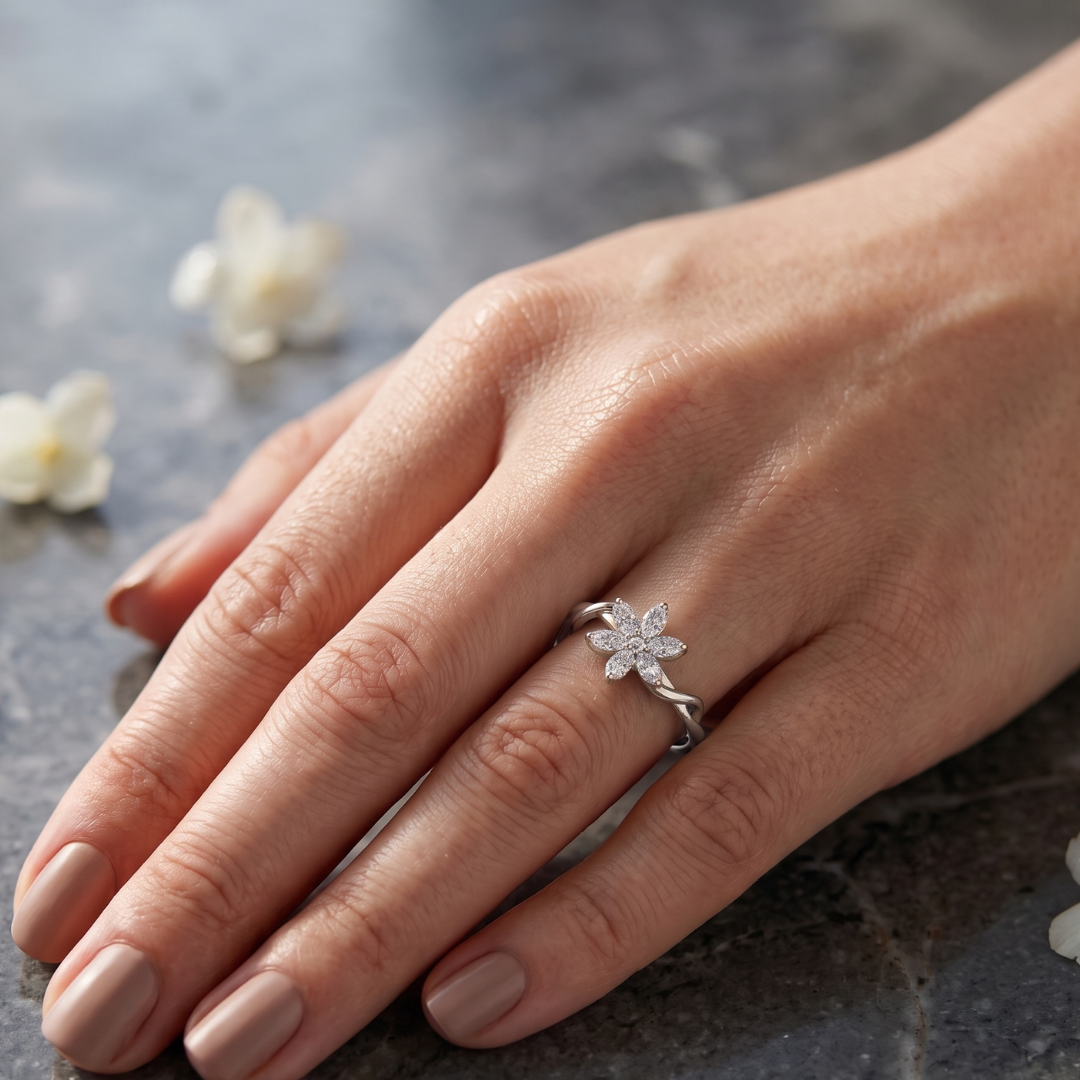 A silver ring shaped like a five-petal flower, set with small clear stones, worn on a woman's hand resting on a dark textured surface with blurred white flowers in the background.