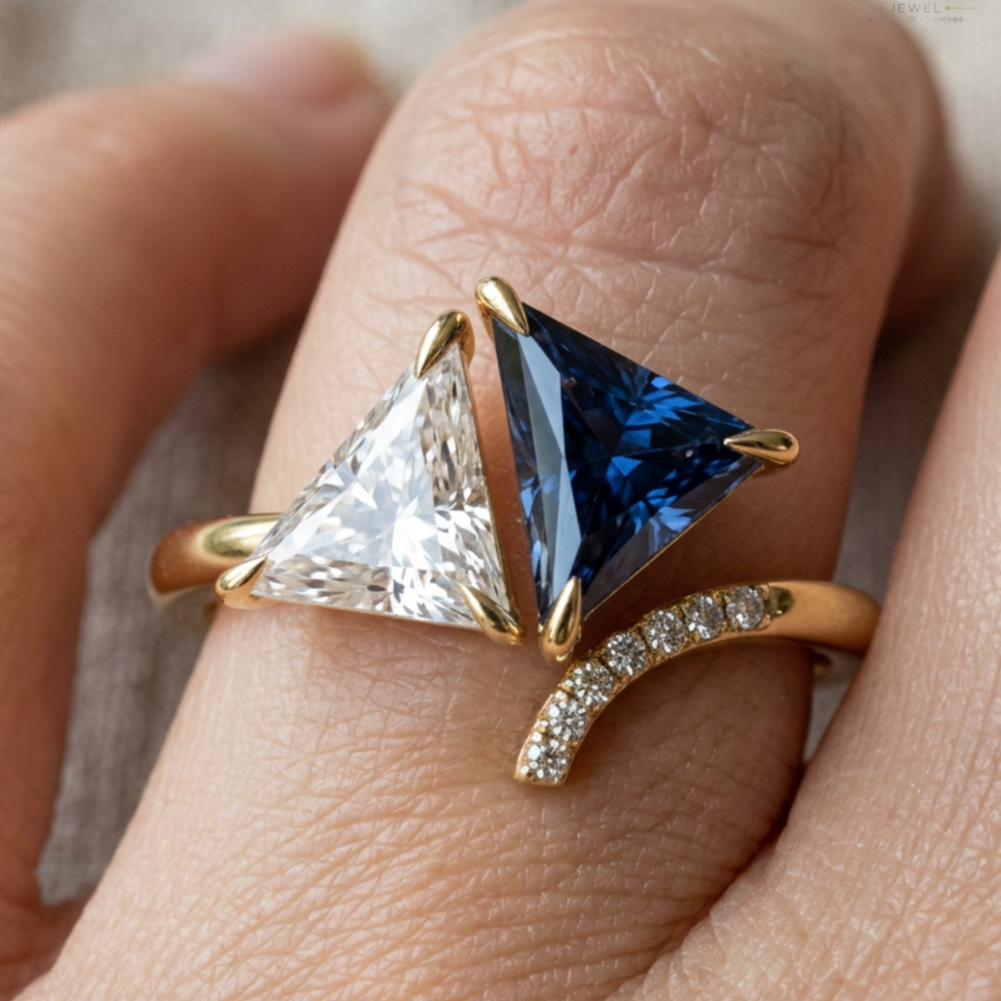A close-up of a gold ring worn on a finger, featuring two triangular gemstones—one clear and one deep blue—along with a curved gold band set with small clear stones.