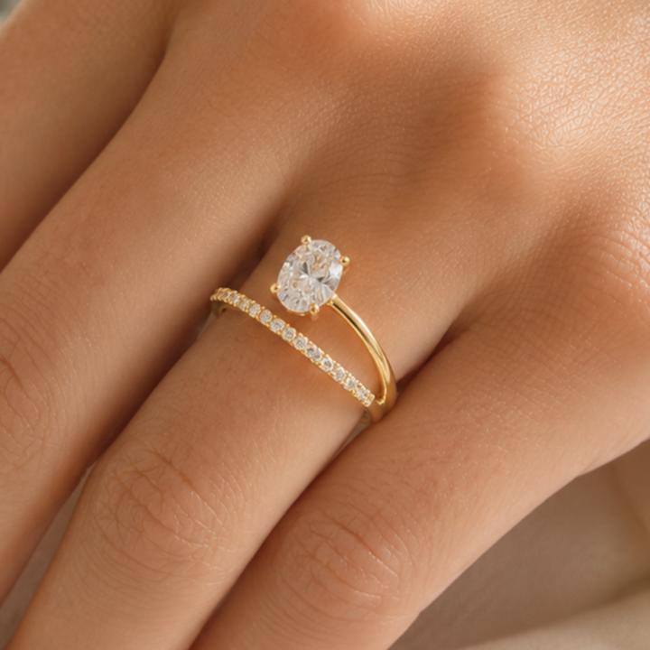 A close-up of a gold ring with a large oval clear gemstone and a smaller pave-studded band, worn on a person's finger.