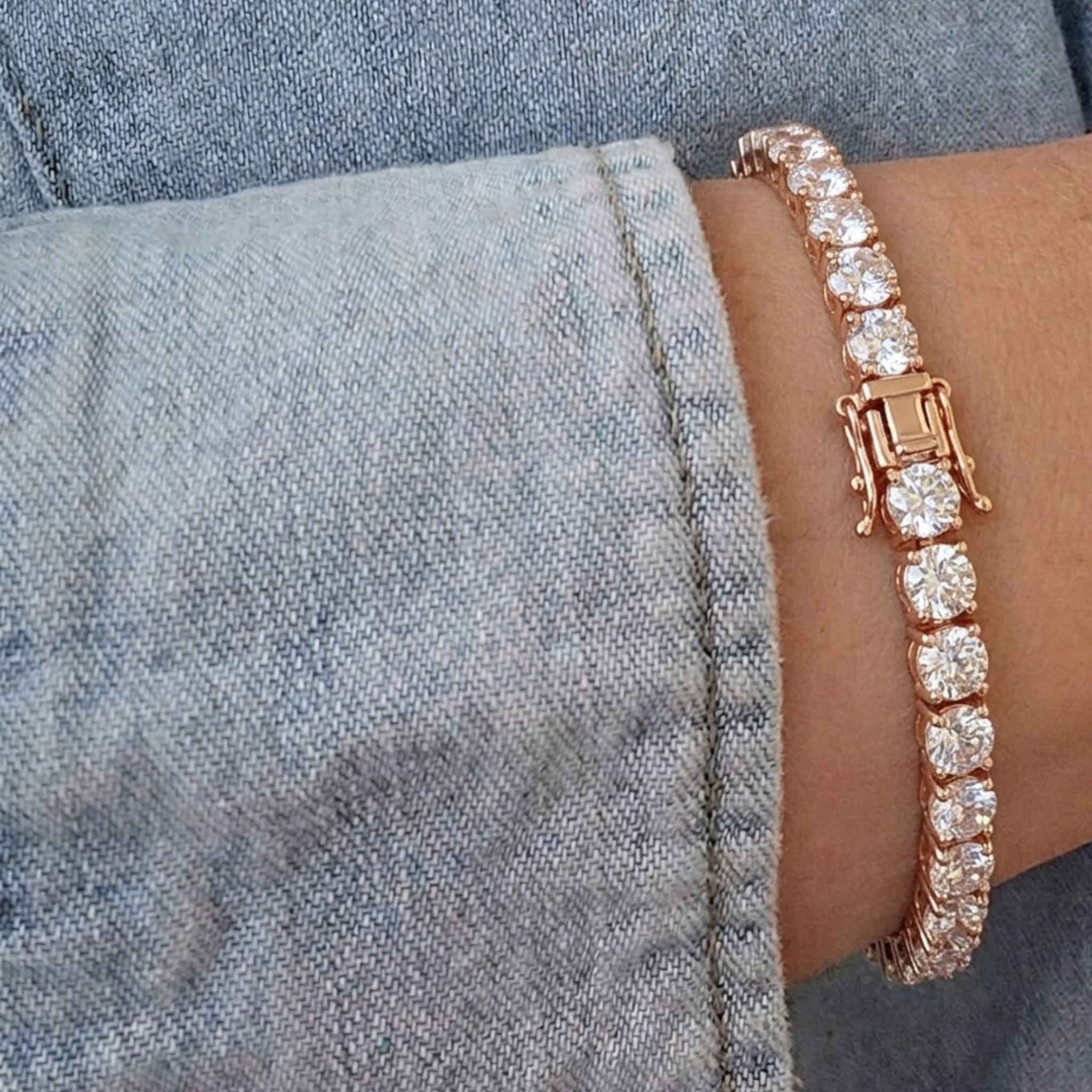 A rose gold tennis bracelet with clear round-cut stones, shown worn on a wrist next to a denim jacket cuff.
