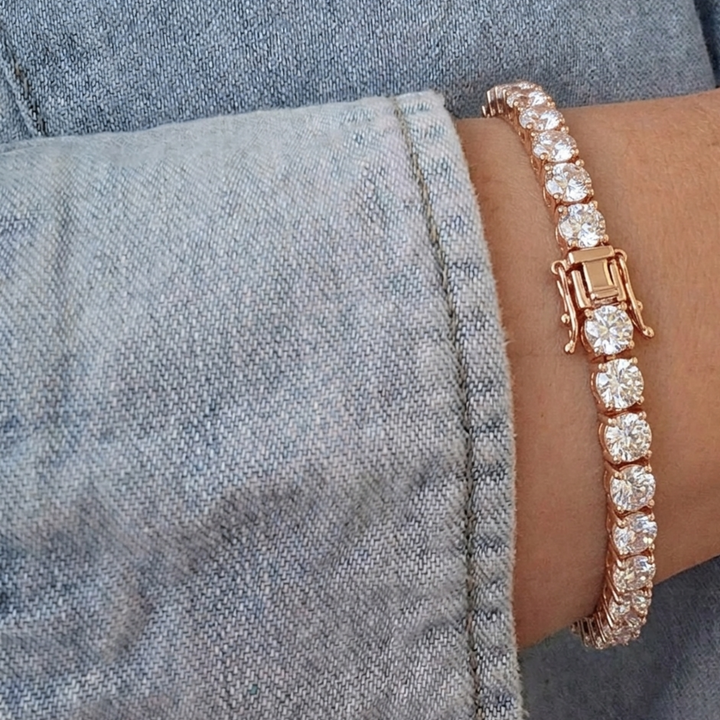 A rose gold tennis bracelet with clear round-cut stones, shown worn on a wrist next to a denim jacket cuff.
