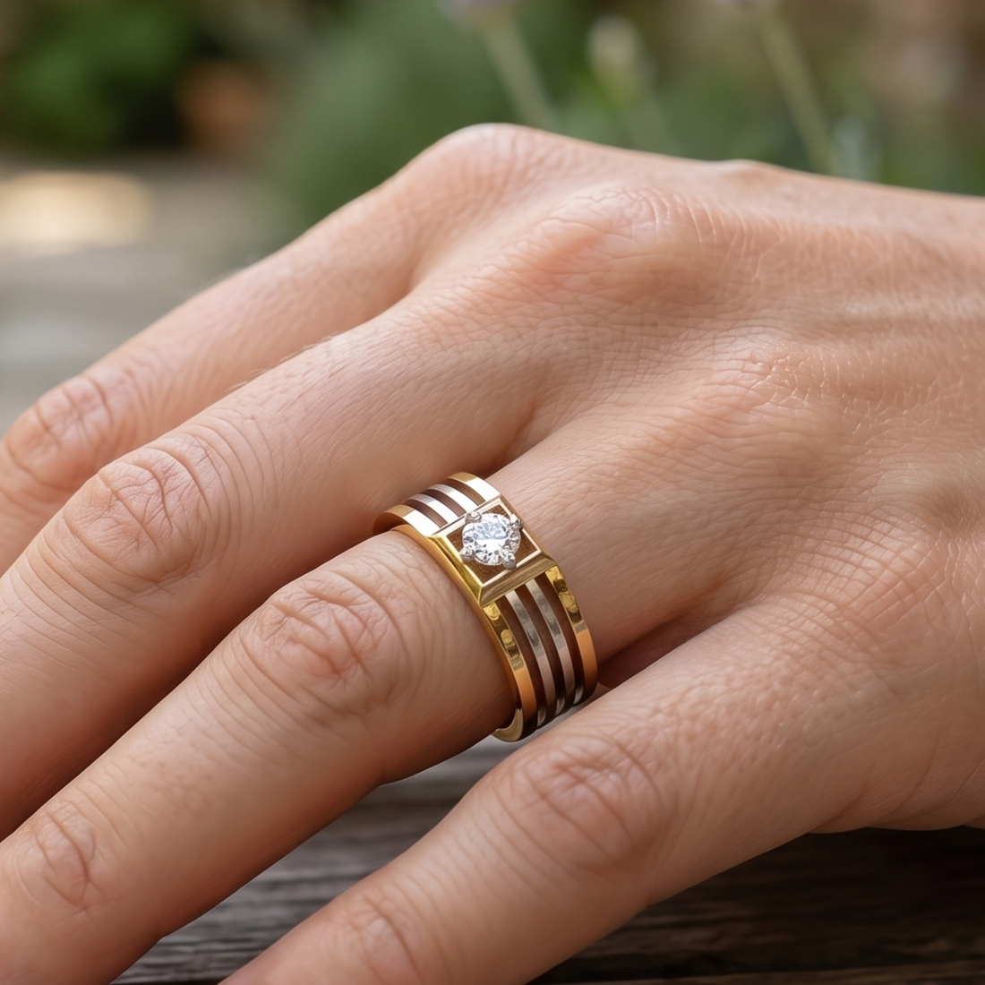 A close-up of a hand wearing a gold ring with three parallel horizontal bands and a central clear gemstone, set against a softly blurred outdoor background.