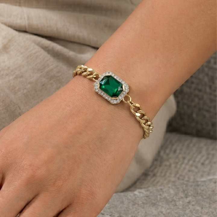 A gold chain bracelet with a central square green gemstone surrounded by a halo of small clear stones, worn on a person's wrist.