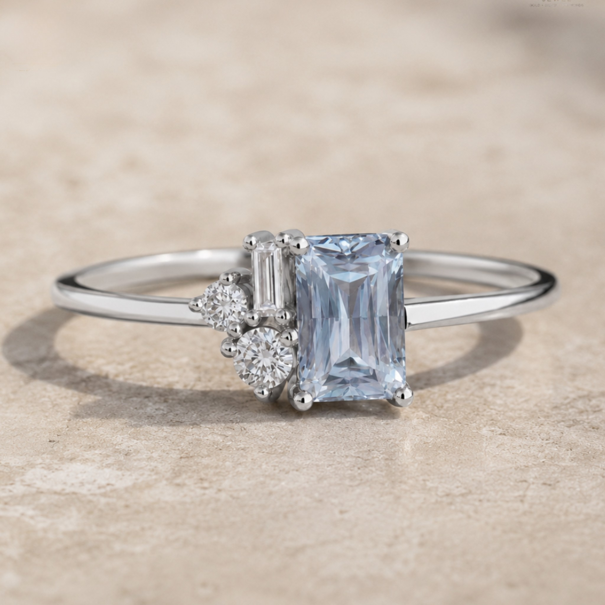 A silver ring featuring a rectangular emerald-cut light blue sapphire center stone, accented by a baguette and round-cut diamonds, resting on a textured beige stone surface.