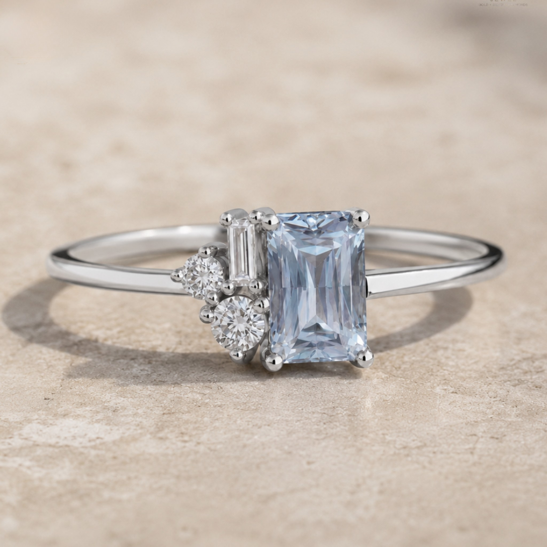 A silver ring featuring a rectangular emerald-cut light blue sapphire center stone, accented by a baguette and round-cut diamonds, resting on a textured beige stone surface.