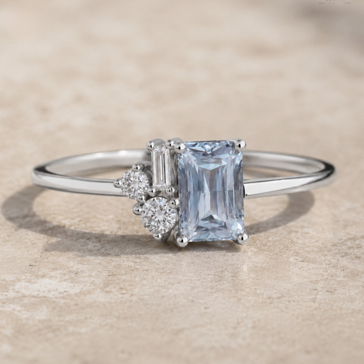 A silver ring featuring a rectangular emerald-cut light blue sapphire center stone, accented by a baguette and round-cut diamonds, resting on a textured beige stone surface.