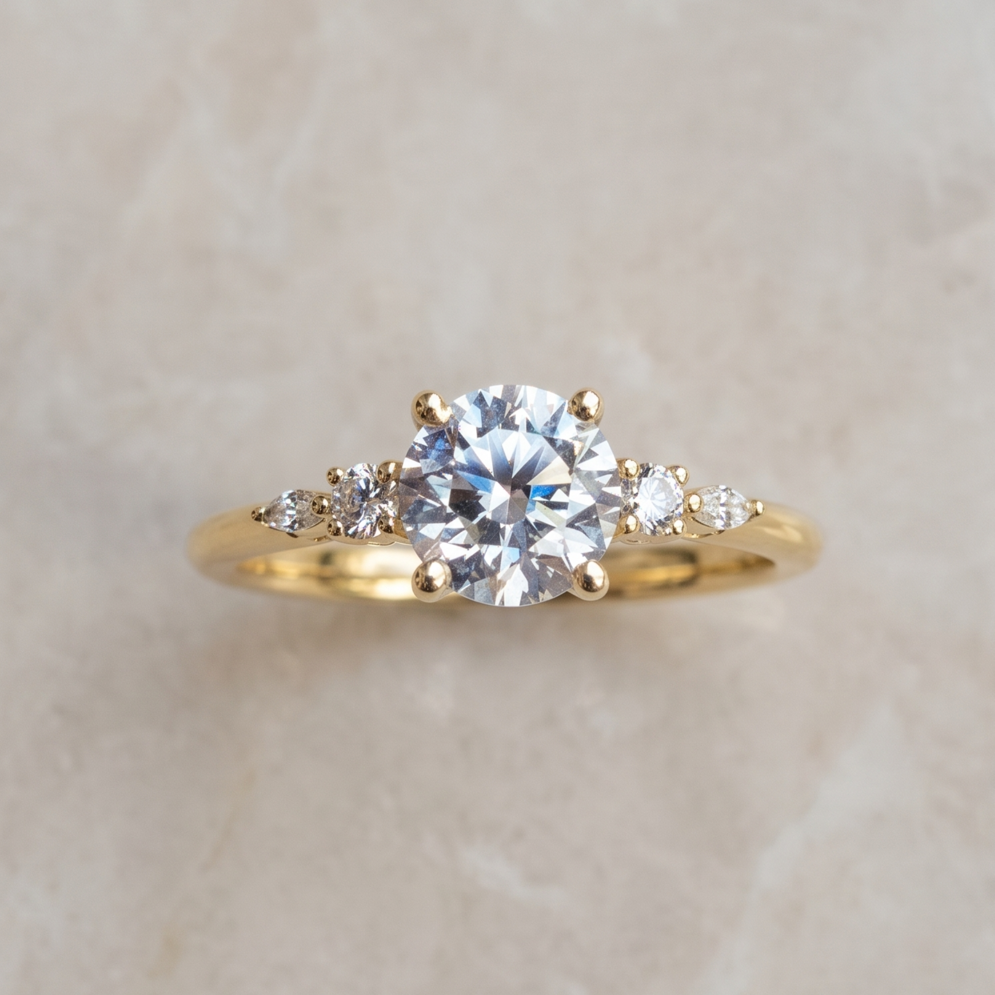 A yellow gold solitaire engagement ring with a round brilliant-cut center stone and smaller accent stones on the shoulders, displayed on a light beige stone background.