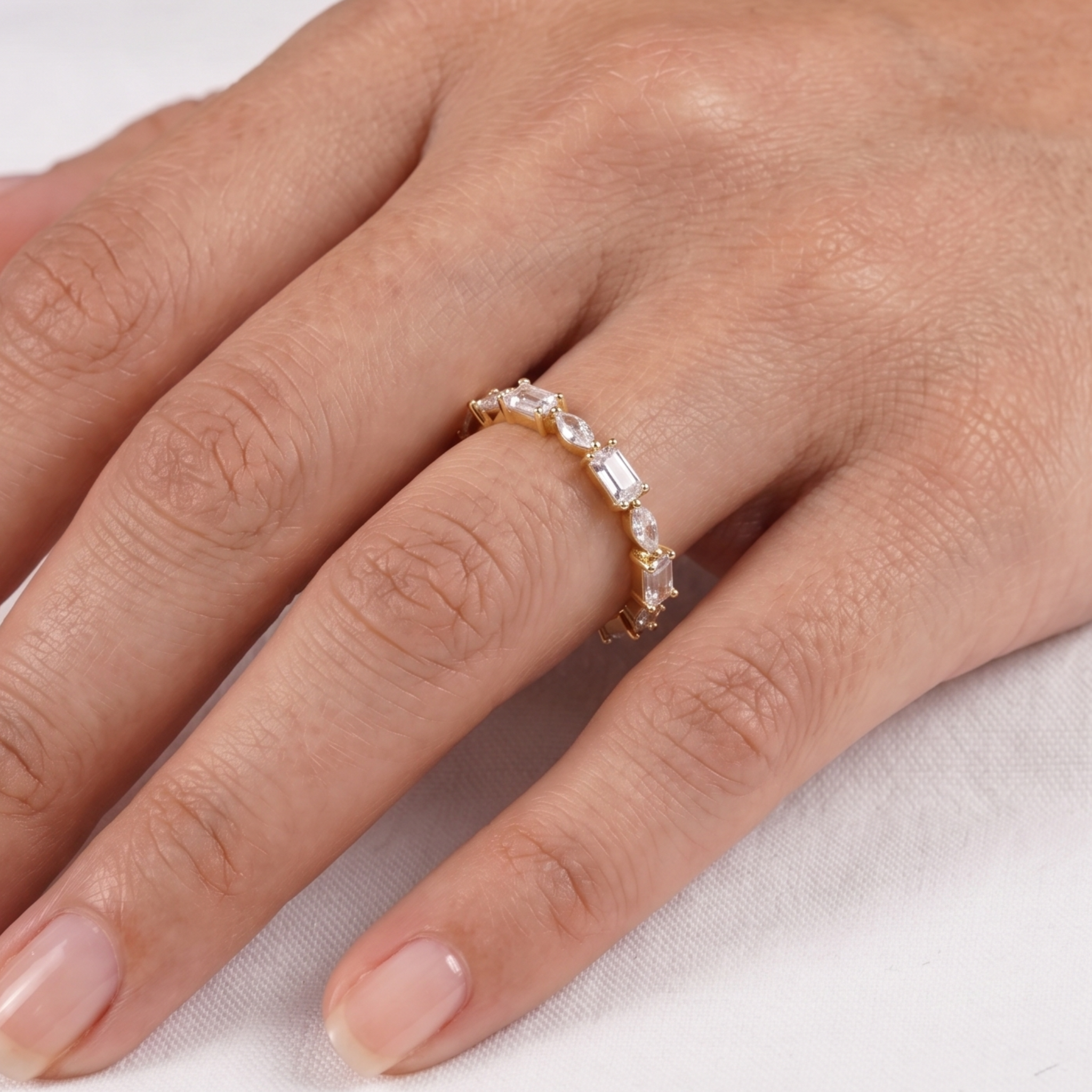 A close-up of a hand wearing a delicate gold eternity ring set with clear baguette-cut gemstones, shown against a soft white background.