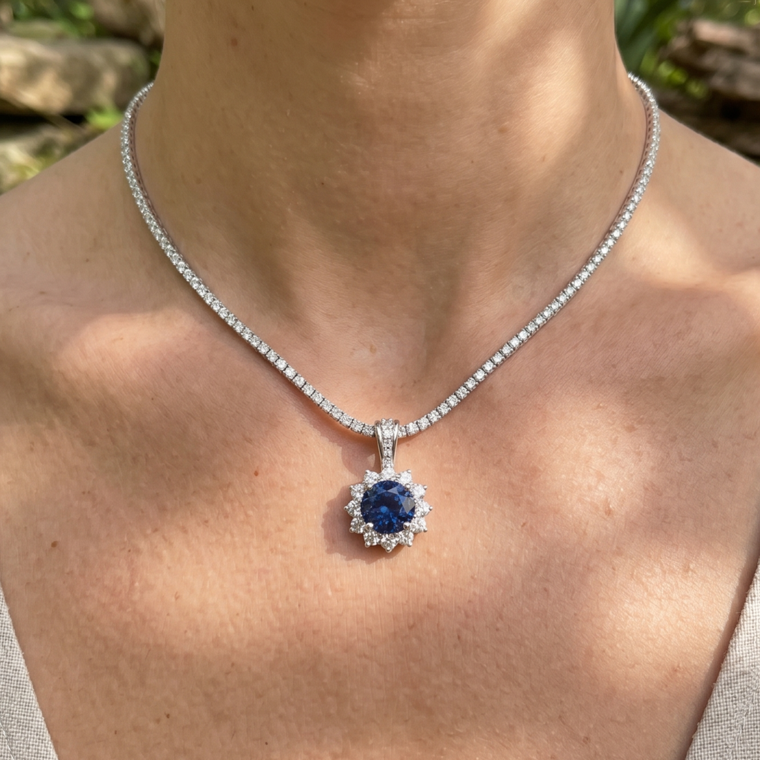 A close-up of a person's neck and chest wearing a silver-toned necklace with a round blue sapphire pendant surrounded by small clear stones.