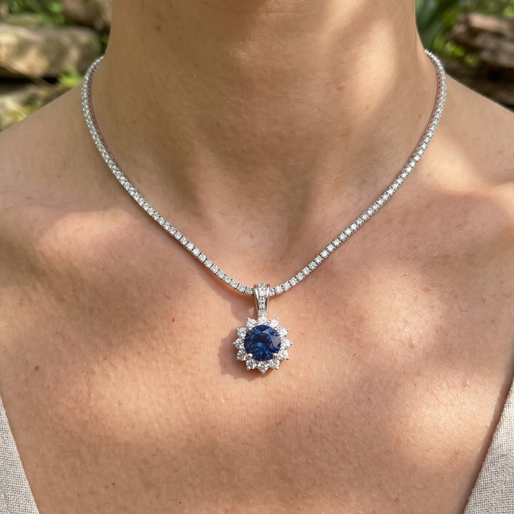 A close-up of a person's neck and chest wearing a silver-toned necklace with a round blue sapphire pendant surrounded by small clear stones.