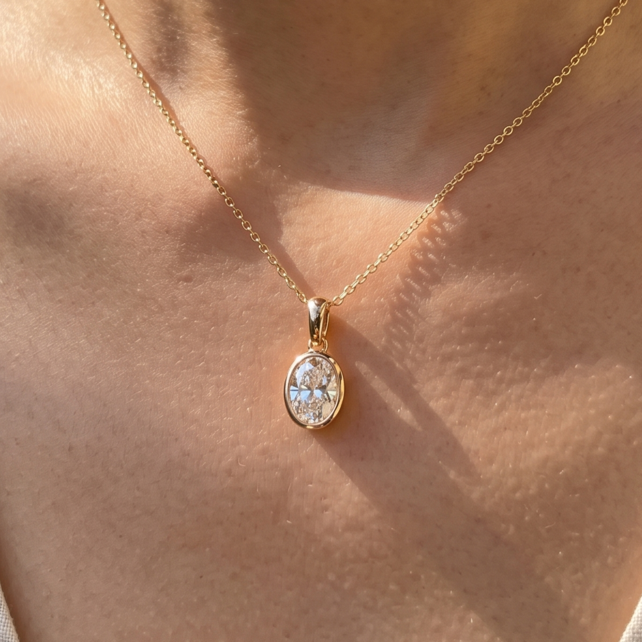 A close-up of a person's neck wearing a delicate gold chain necklace with an oval pendant set with clear stones, shown in warm natural sunlight.