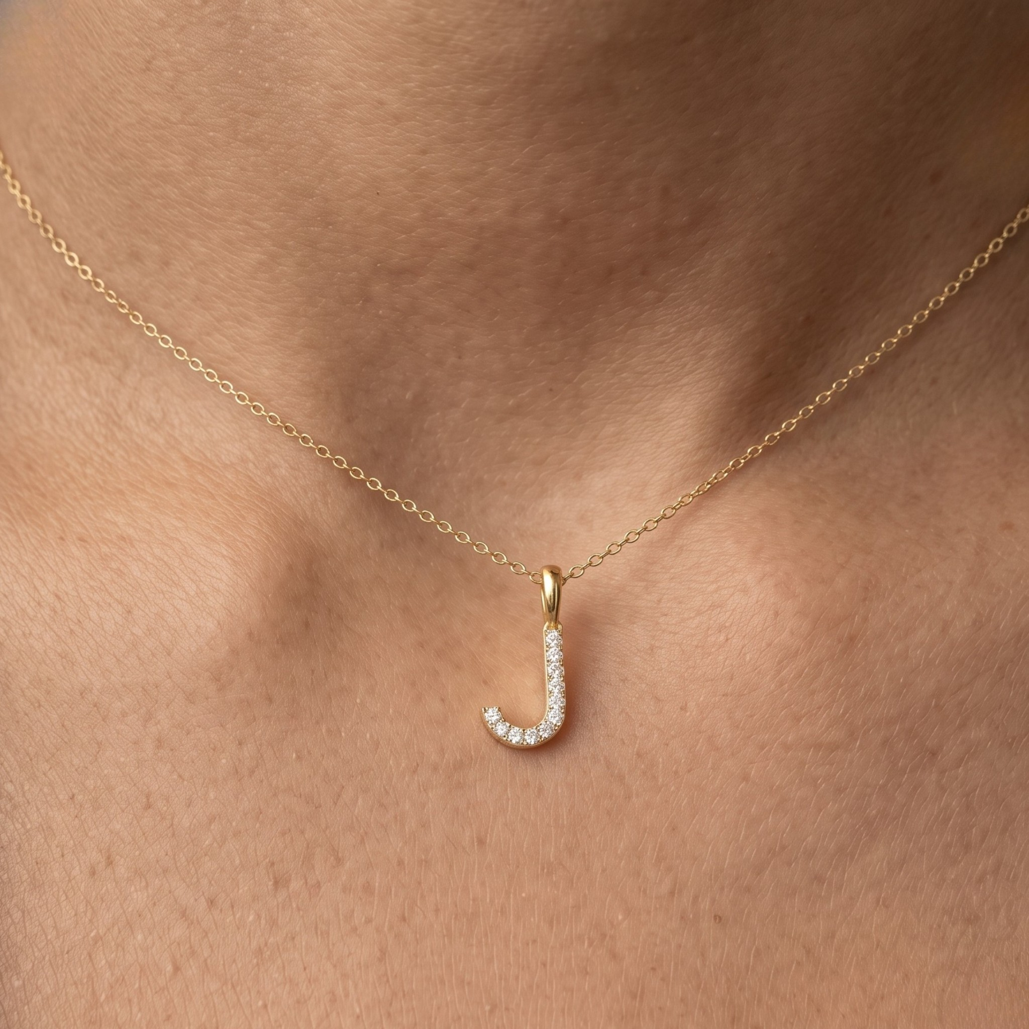 A close-up of a person's neck wearing a delicate gold chain necklace with a small curved initial pendant paved with clear stones.