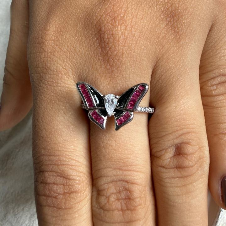 A silver butterfly ring with black enamel and pink gemstone accents, worn on a finger.