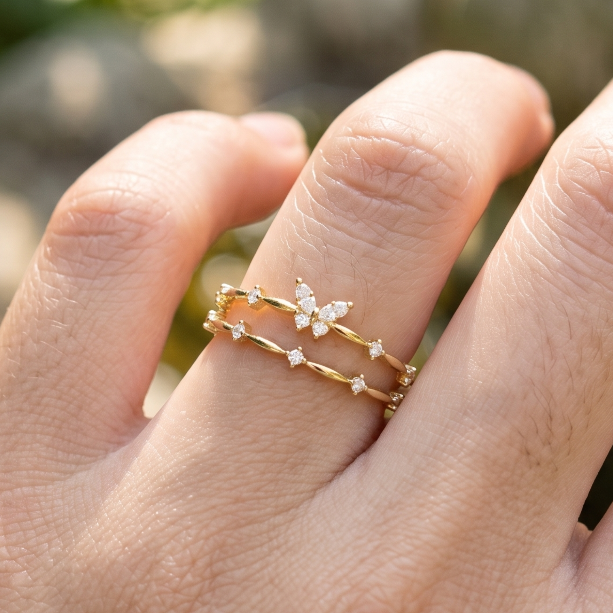 A close-up of a hand wearing two delicate gold rings. One ring features a small gold butterfly embellished with clear stones, and the other is a thin gold band with small star-like clear stone accents.