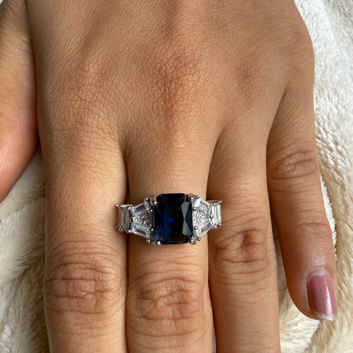 A close-up of a hand wearing a silver ring with a rectangular dark blue sapphire center stone and baguette-cut side stones, resting on a white textured blanket.