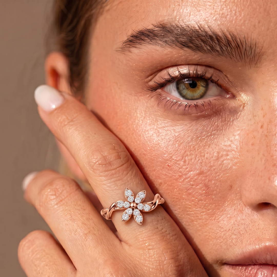 A close-up lifestyle shot of a model wearing a rose gold flower ring with clear gemstones on her finger, with her hand resting against her cheek.