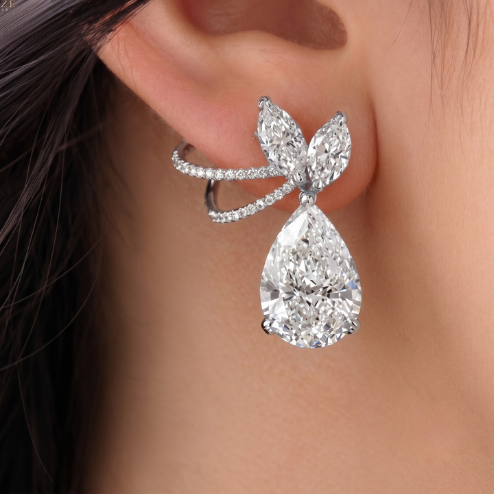 A close-up of a model's ear wearing a silver-toned earring with a large pear-cut clear gemstone and smaller marquise-cut stones, accented by tiny pavé stones along a curved band.