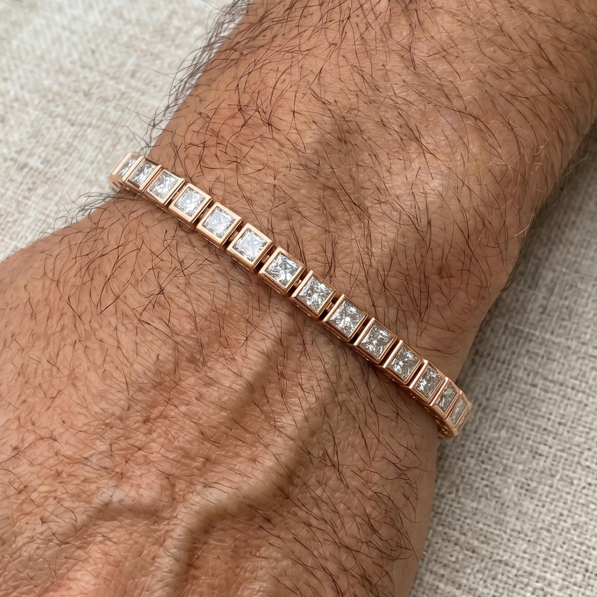 A close-up of a rose gold tennis bracelet with square-cut clear stones, worn on a hairy wrist against a neutral textured fabric background.